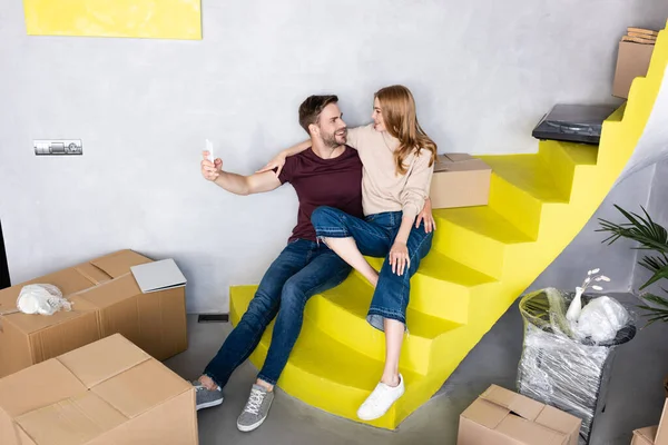 Couple joyeux assis sur des escaliers jaunes et l'homme prenant selfie près des boîtes en carton — Photo de stock