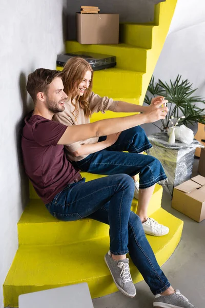 Alegre pareja sentado en amarillo escaleras y tomando selfie cerca de cajas de cartón — Stock Photo