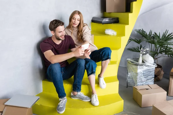 Pareja sentada en las escaleras y mirando el teléfono inteligente cerca de cajas, concepto de reubicación - foto de stock