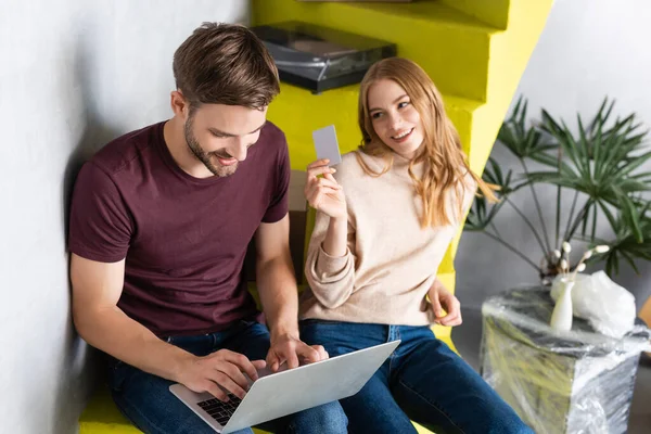 Young woman holding credit card and looking at boyfriend using laptop — Stock Photo
