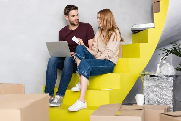 Selective focus of man with laptop looking at girlfriend holding credit card while sitting on stairs — Stock Photo