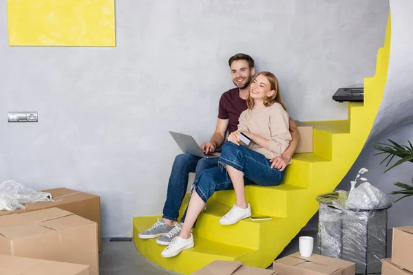 Homem sentado em escadas perto de mulher com cartão de crédito perto de laptop e caixas — Fotografia de Stock