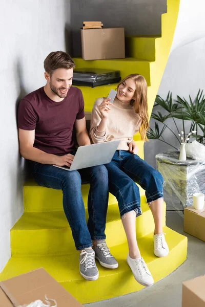 Mujer sosteniendo la tarjeta de crédito y mirando al novio usando el ordenador portátil y sentado en las escaleras - foto de stock