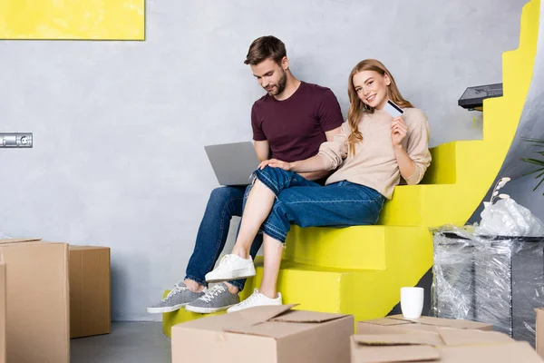 Mujer joven sosteniendo la tarjeta de crédito cerca del hombre usando el ordenador portátil y sentado en las escaleras amarillas, concepto de reubicación - foto de stock