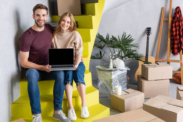 Pareja que sostiene el ordenador portátil con la pantalla en blanco mientras está sentado en escaleras cerca de cajas, concepto de reubicación - foto de stock