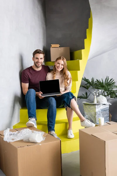 Alegre pareja sosteniendo portátil con pantalla en blanco mientras está sentado en las escaleras cerca de cajas, concepto de reubicación - foto de stock