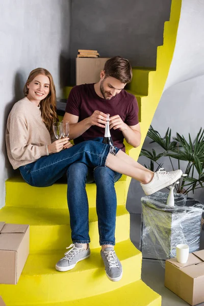 Mann öffnet Flasche, während freudige Frau Gläser hält und auf Treppe sitzt, bewegtes Konzept — Stockfoto