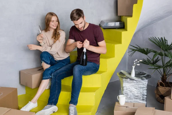 Hombre abriendo botella mientras mujer joven sosteniendo gafas y sentado en escaleras amarillas, concepto en movimiento - foto de stock