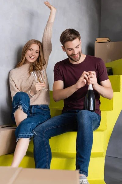 Hombre apertura botella mientras complacido joven mujer sosteniendo gafas y sentado en escaleras, concepto en movimiento — Stock Photo