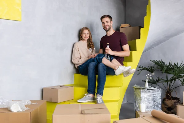 Hombre apertura botella mientras complacido joven mujer sosteniendo gafas y sentado en escaleras, concepto en movimiento - foto de stock