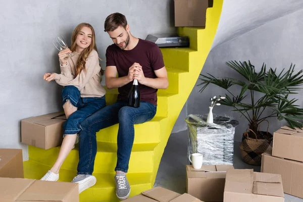 Man opening bottle while pleased girlfriend holding glasses and sitting on stairs, moving concept — Stock Photo