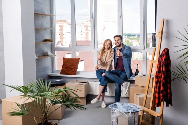 Pleased boyfriend and girlfriend holding glasses with champagne near carton box, moving concept — Stock Photo