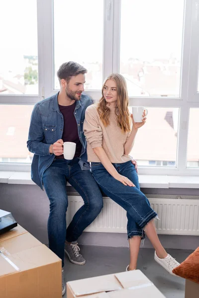 Pareja complacida sosteniendo tazas con té cerca de cajas, concepto móvil - foto de stock