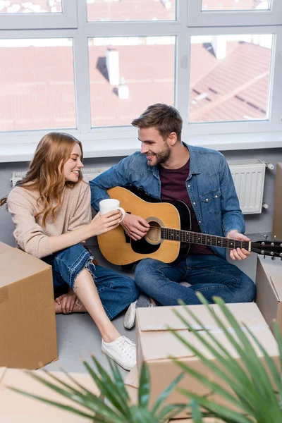 Enfoque selectivo de hombre joven tocando la guitarra acústica cerca de la mujer con la taza y cajas de cartón - foto de stock