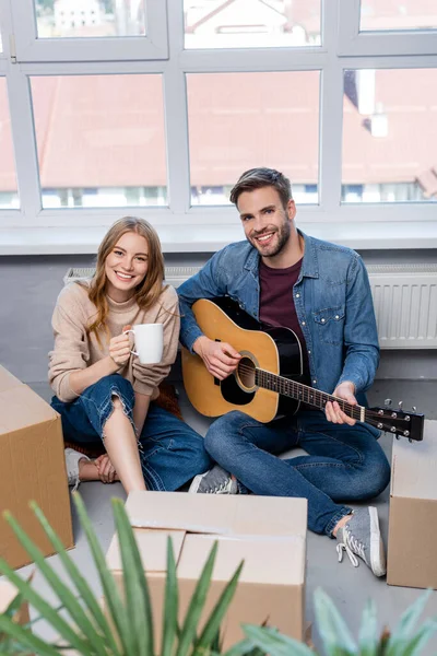 Foco seletivo do jovem tocando guitarra acústica perto de mulher alegre com caixas de copo e caixa — Fotografia de Stock