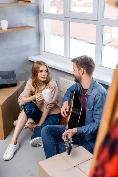 Vista de ángulo alto de hombre joven tocando la guitarra acústica cerca de la mujer con la taza y cajas de cartón - foto de stock