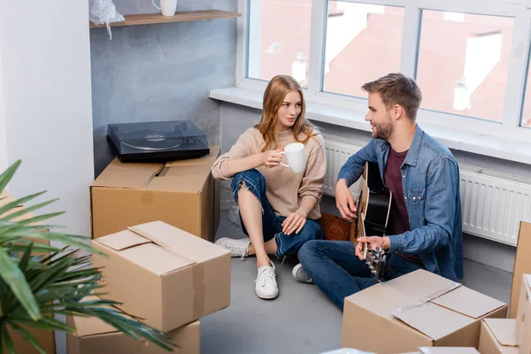 Foco seletivo do jovem sentado no chão tocando guitarra acústica perto de mulher com copo e caixas de papelão — Fotografia de Stock