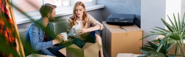 Selective focus of joyful woman holding cup while unpacking box with boyfriend, panoramic shot — Stock Photo