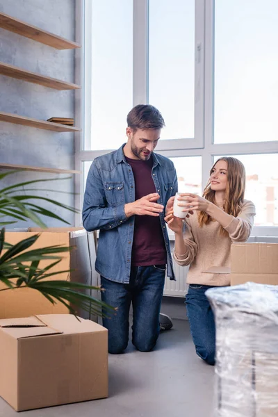 Homme regardant la tasse dans les mains de la femme tout en déballant les boîtes en carton dans la nouvelle maison, concept mobile — Photo de stock