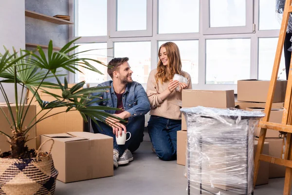 Homme et femme tenant des tasses et se regardant tout en déballant des boîtes en carton dans une nouvelle maison — Photo de stock