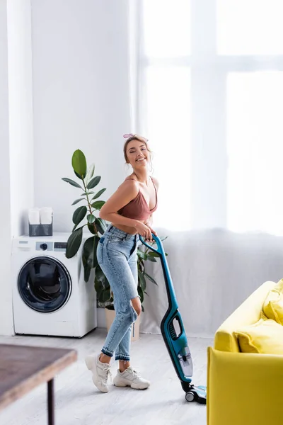 Dona de casa moderna com aspirador de pó olhando para a câmera perto de planta e máquina de lavar roupa — Fotografia de Stock