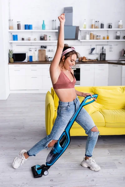 Возбужденная домохозяйка танцует с пылесосом возле желтого дивана — стоковое фото