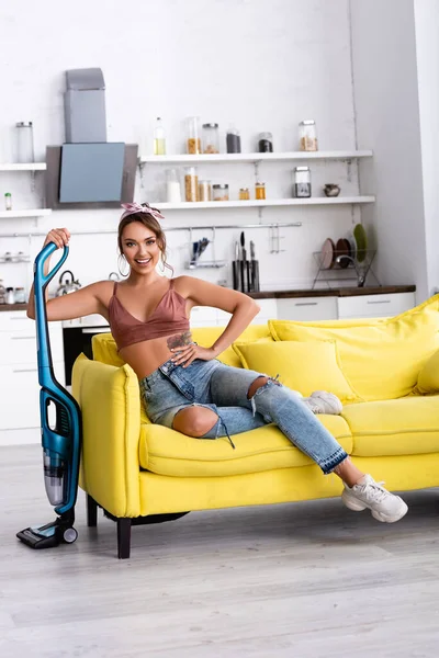 Femme au foyer avec la main sur la hanche assis sur le canapé et tenant aspirateur à la maison — Photo de stock