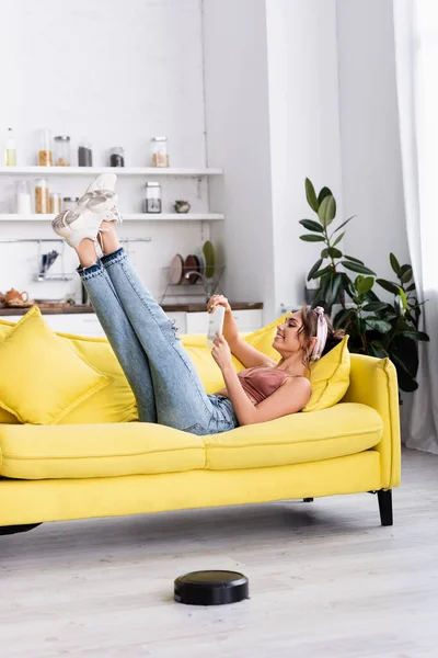 Woman with legs in air lying on couch near robotic vacuum cleaner at home — Stock Photo