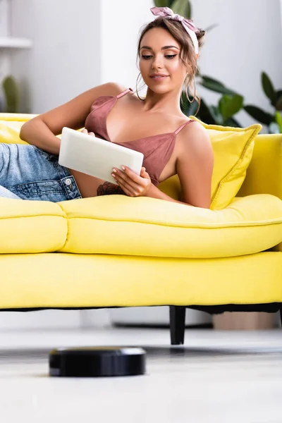 Concentration sélective de la femme en utilisant une tablette numérique sur le canapé et l'aspirateur robotique — Photo de stock