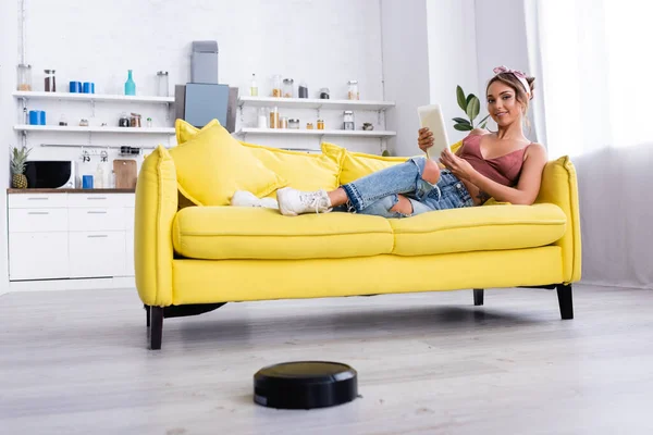 Женщина лежит на диване с цифровым планшетом и роботизированным пылесосом на полу — стоковое фото