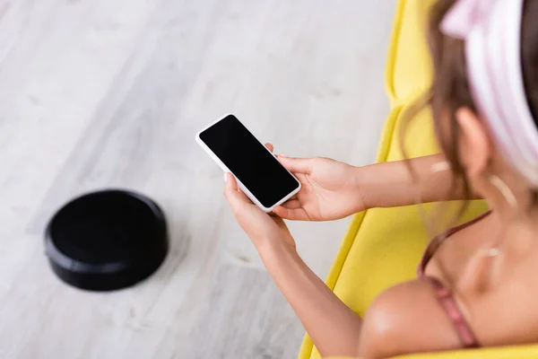 Enfoque selectivo de ama de casa utilizando el teléfono inteligente mientras está sentado en el sofá cerca de la aspiradora robótica - foto de stock