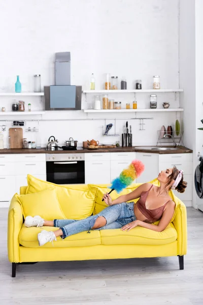 Young housewife blowing on dust brush while lying on couch at home — Stock Photo