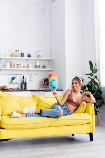 Молодая домохозяйка держит пыльную щетку, лежа дома на диване — стоковое фото