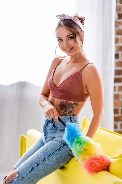 Selective focus of tattooed housewife looking at camera while holding dust brush near couch — Stock Photo