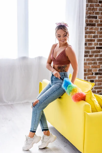 Молода домогосподарка в джинсах прибирає диван з пиловим пензлем — стокове фото