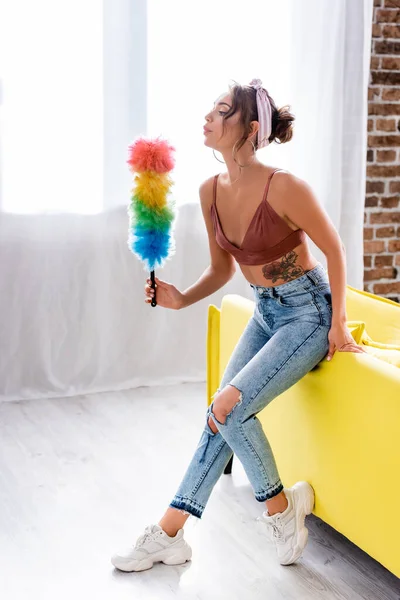 Modern housewife blowing on dust brush near yellow sofa at home — Stock Photo