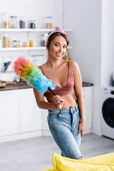 Foco seletivo de jovem dona de casa olhando para a câmera e segurando escova de poeira em casa — Fotografia de Stock