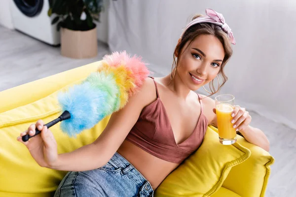 Selektiver Fokus der Hausfrau mit Staubpinsel und einem Glas Orangensaft auf der Couch — Stockfoto