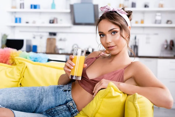 Enfoque selectivo de la mujer joven con vaso de jugo de naranja mirando a la cámara cerca del cepillo de polvo en el sofá - foto de stock