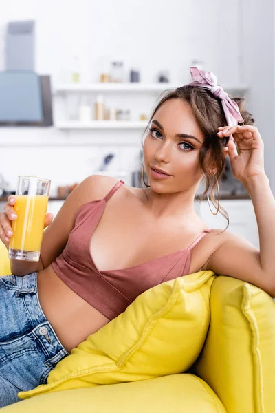 Молода жінка дивиться на камеру, тримаючи склянку апельсинового соку на дивані — стокове фото