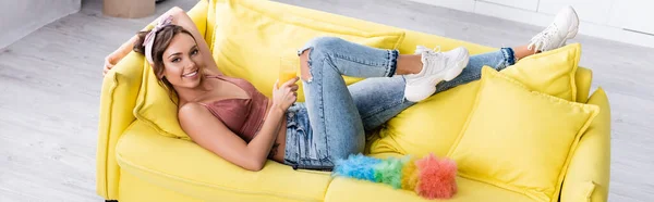 Foto panoramica di giovane casalinga che guarda la macchina fotografica mentre tiene in mano un bicchiere di succo d'arancia vicino alla spazzola della polvere sul divano — Foto stock