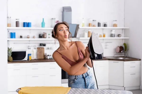 Femme au foyer choquée tenant du fer près du t-shirt sur une planche à repasser — Photo de stock