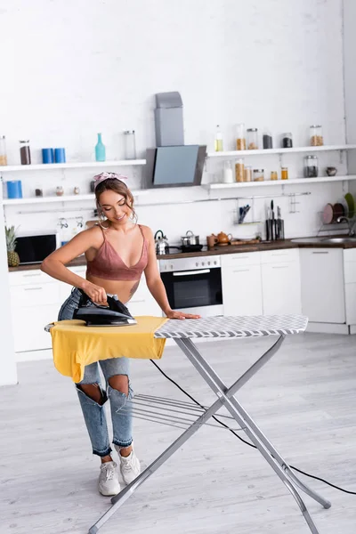 Jeune femme au foyer repassage t-shirt sur planche à repasser dans la cuisine — Photo de stock