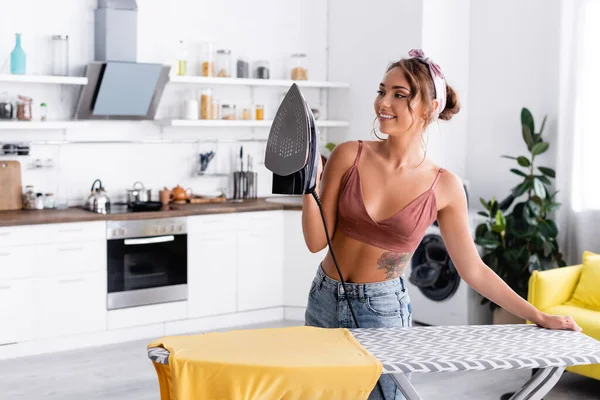 Femme au foyer tatouée avec bandeau tenant du fer près du t-shirt sur planche à repasser dans la cuisine — Photo de stock