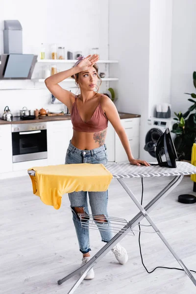 Müde Hausfrau steht neben Bügeleisen und T-Shirt auf Bügelbrett — Stockfoto