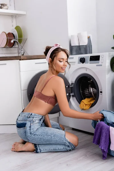 Pieds nus femme au foyer assis sur le sol près des vêtements et machine à laver dans la cuisine — Photo de stock