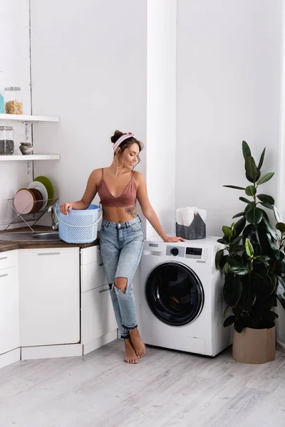 Descalzo ama de casa sosteniendo cesta de lavandería cerca de la lavadora en la cocina - foto de stock