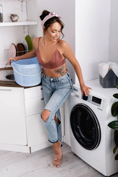 Brunette woman in headband touching washing machine and holding laundry basket in kitchen — Stock Photo