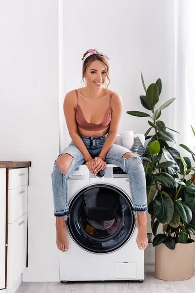 Barfüßige Hausfrau blickt in die Kamera, während sie auf der Waschmaschine sitzt — Stockfoto