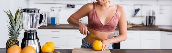 Панорамний урожай молодої жінки, що ріже апельсин біля блендера на кухні — стокове фото